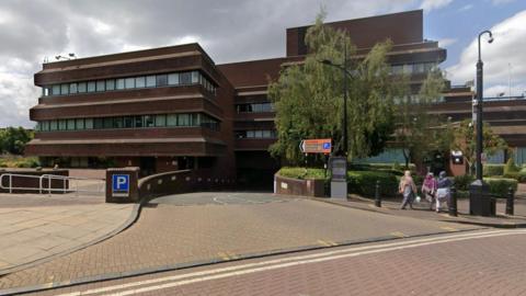 Dark brick building with glass windows with a road leading down to parking underneath the building. A blue P sign on the left hand side and three women walking away from the building on the right hand side.