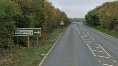 The A134 in Stoke Ferry. It is a single carriageway road and there is a green road sign on the left of the picture which says Stoke Ferry and is directing people to it on the right.