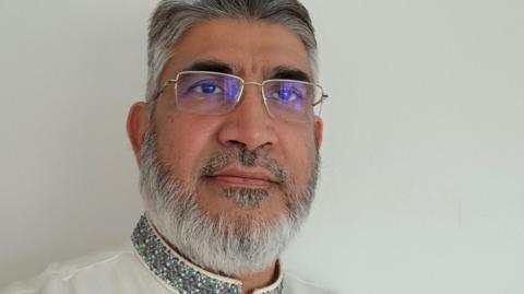 Tofail Awan, who has greying hair and a beard, is wearing silver-rimmed glasses. He is wearing a white-collared thobe with silver embroidery.