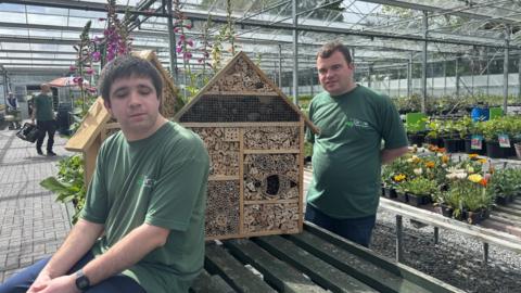 Two men in green shirts stood side by side next to a wooden bug hotel ina greenhouse.
