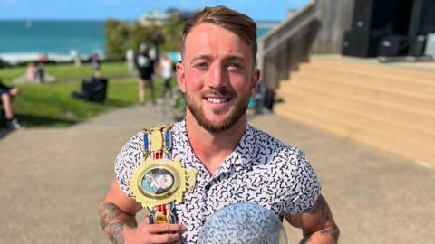 Brad Pauls is smiling at the camera with the sea behind him and his golden title belt draped over his right shoulder