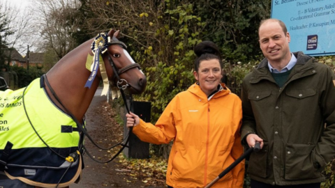 Emma Webb, who is holding the reins of resin horse Max, is wearing an orange raincoat and his standing next to Prince William, who is wearing dark green waxed coat