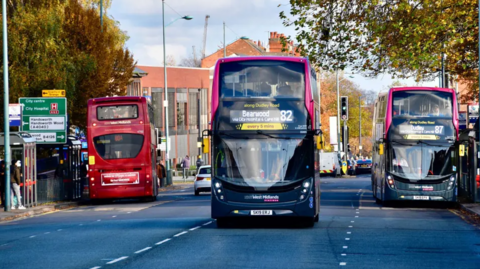 Three buses can be seen on a road travelling in different directions. One is red and two others are dark blue and pink.