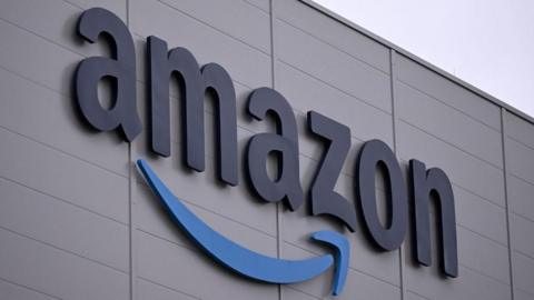 The Amazon logo, which is the world 'Amazon' with its signature stylised arrow underneath, on the side of a grey building with a light overcast sky in the background