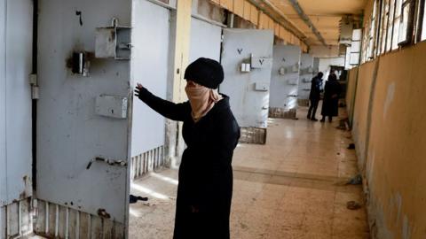 A woman opens a prison door in a corridor of Saydnaya prison.  She is wearing a dark coat and hat and her face is covered with a scarf.  In the background, along the corridor, is another group of people.