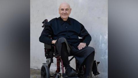 A smiling Bill Ward in black shirt, trousers and shoes, sits in a wheelchair against a plain grey background