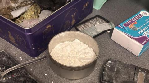A saucepan of powder, a box of prescription drugs and a cake tin of drugs can be seen on a kitchen work bench.