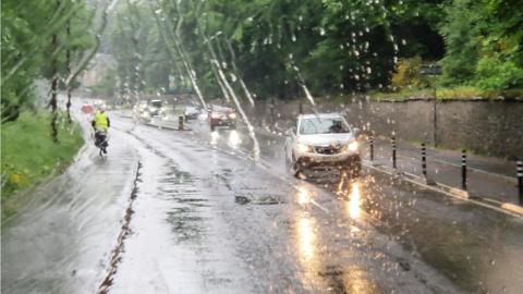 Rain falling across a road with oncoming traffic driving on wet roads and trees on either side