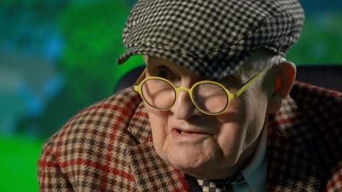 David Hockney wearing a flat cap and round yellow glasses is looking into a camera and talking.