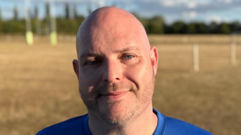 Matthew Pearson smiles straight at the camera while standing on a rugby pitch.