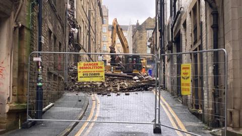 A yellow mechanical digger behind metal fencing lifting debris.