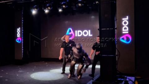 Two men dressed in black rush towards a grey robot toppling forward.on stage as it topp