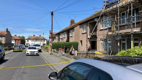 An image of the residential street where the incident happened. Yellow police tape has blocked off the house involved. A police car can be seen behind the tape and two officers are standing in front of the property. 