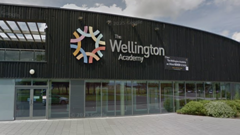Google maps street view of Wellington Academy. The building is wide and covered in black wooden panels, with a large Wellington Academy logo on the front.