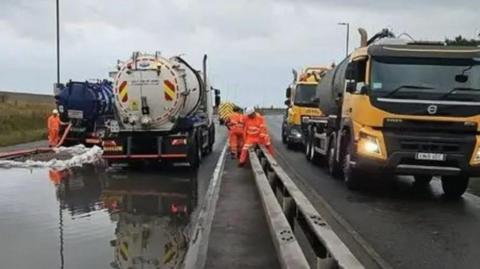 Two tankers on a road, with the left side flooded and water is being drained, while workers wearing orange overalls carry out work. There are also two other lorries on the right.