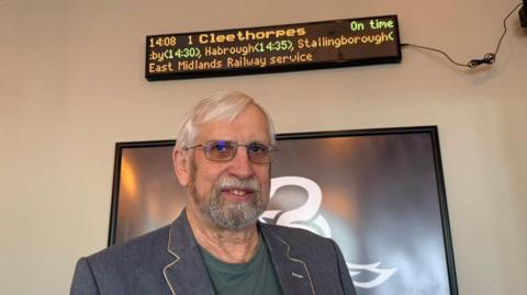 The photo shows a man smiling at the camera. He's wearing a blue blazer, a green top and glasses. He's standing in front of a black frame. Above his head is a train departure board, which reads "Cleethorpes" and various other railway stations in LED lighting. 