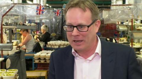 Mike Raybould, wearing a pale-coloured shirt and a black suit jacket, is stood in a pottery factory with pottery products and Union Jack bunting behind him.