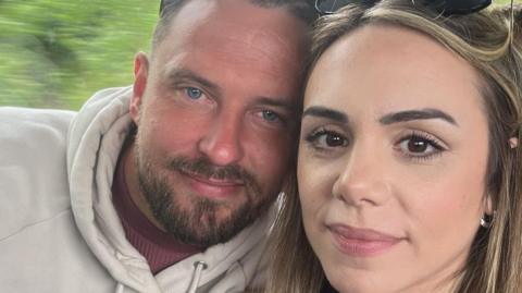 Jason and Mimi take a selfie while sat on a train, their heads touching.