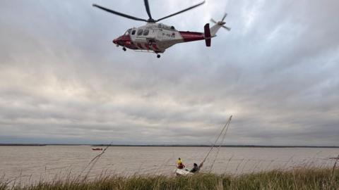 A red and white helicopter flying in the sky, over water, with two people in a white boat that is listing. Another red vessel is in the water, and there are reeds in front of the water. A cable is going from the helicopter to the boat.