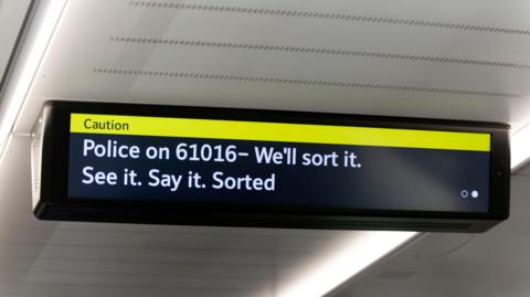 An electronic display inside a London Underground train showing a yellow “Caution” banner and a message advising passengers to contact police on 61016, alongside the slogan “See it. Say it. Sorted”.
