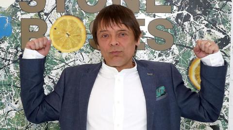 Mani with both arms raise standing in front of a large image of the Stone Roses' debut album artwork