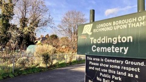 A sign for Teddington Cemetery in the London Borough of Richmond upon Thames beside a path, with fencing, trees and allotment plots visible in the background.