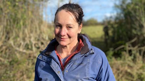 A woman wearing a blue jacket smiling at the camera with a hedgerow behind her