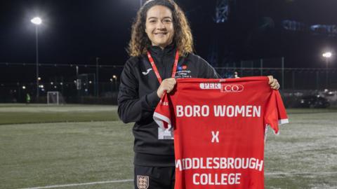 Sammie Leigh is dressed in black football kit and she holds footballs and a t-shirt reading 'BORO WOMEN X MIDDLESBROUGH COLLEGE'. She is standing on a football pitch at night. 