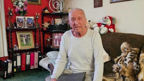 An older man wears a light grey long-sleeved top and sits on a sofa holding a dark file or binder. Behind him are shelves containing framed family photographs, flowers, ornaments and ring binders. A patterned throw covers part of the sofa and soft furnishings fill the room.