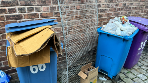 Blue bins overflowing with cardboard in an alley in Liverpool. Trays made of wire mesh and other cardboard boxes are strewn around on the cobbles next to the bins. 