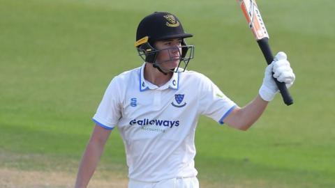 Charlie Tear in white Sussex kit and blue helmet raises the bat to acknowledge the crowd