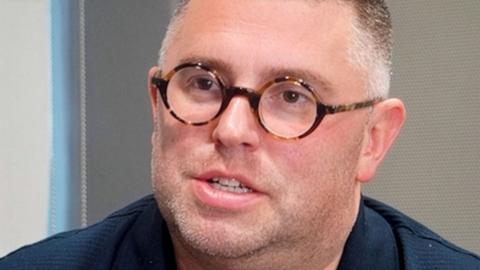A close up of Alex Wood, who is wearing a navy blue shirt and round glasses. 