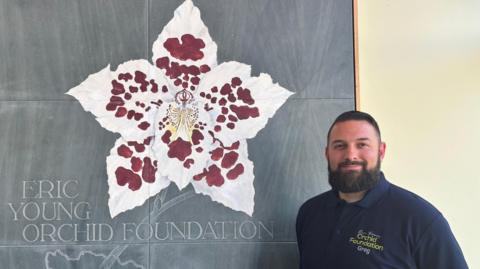 Greg Griffis is smiling as he stands next to a large slate sign. There is a red and white orchid flower painted on it and an outline of Jersey etched into it with the words ERIC YOUNG ORCHID FOUNDATION.