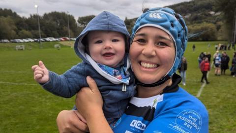 Trish Brown in a blue rugby shirt and blue helmet smiling, holding her baby son, who is also smiling and wearing a blue hoody. They are on a rugby pitch.