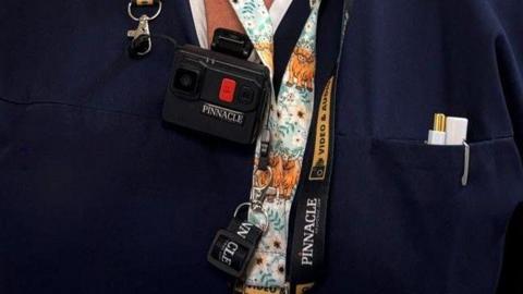 An NHS worker has a lanyard on reading 'Pinnacle' and a black body camera on. They have two white pens in their uniform pocket.