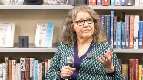 Angela Montgomery holding a microphone and speaking in front of bookshelves filled with various books in a bookstore setting.