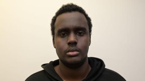 A custody mugshot of Mohamed Mohamoud, who has a serious expression on his face as he stares at the camera. He has short curly black hair and is wearing a black hoodie
