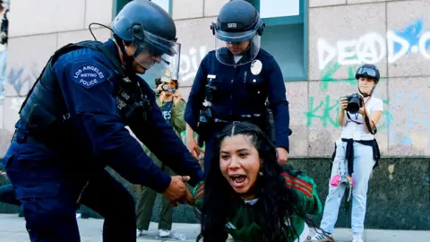 This image shows two police officers from the Los Angeles Metro Police detaining a woman. The officers are wearing protective gear, including helmets and vests. One officer is holding the person's arms while the other stands nearby.

In the background, there is graffiti on the wall, including the word "DEAD" and other illegible markings. Additionally, a photographer wearing a helmet is capturing the scene with a camera. The image appears to depict law enforcement in action during a public disturbance or protest. 