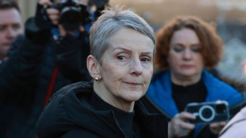 A close-up image of Lady Eleanor Donaldson outside court in Newry. Behind her are reporters with cameras and phones pointed at her. Lady Donaldson is an older woman with short grey hair. She has a neutral expression on her face. She is wearing a large, black coat.