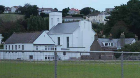 The picture shows a landscape scene featuring a prominent white building with multiple sections and a tower and a grassy field in the foreground, enclosed by a metal fence.