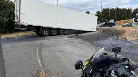 A trailer of a large articulated lorry which has no cab positioned across a roundabout approach. The trailer has grounded with visible damage to the road surface beneath. Several other vehicles are visible in the background, while part of a police motorcycle can be seen in the foreground. There are trees, road signs and dry grass verges surrounding the junction.