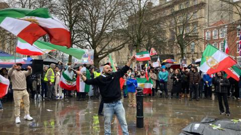 A man is waving the Iranian flag in front of a crowd of people also waving Iranian flags. They are standing in a square which is wet from rain.