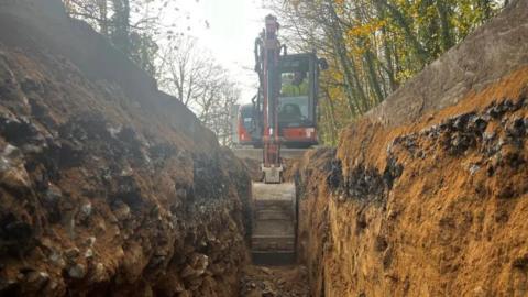 A small digger is digging a channel though sandy and stony earth which is piled up both sides of the deep gully.