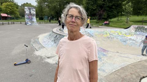 A woman wearing a pink t-shirt stares straight at the camera. She is wearing glasses and her hair is tied back. Behind her is a skate park, including a steep skate ramp. It is covered in grafitti. A blue child's scooter is on the woman's left hand side behind her.