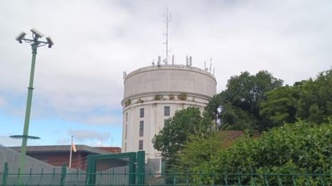 A round, white water tower set in grassland but surrounded by other water works buildings, trees and green metal security fencing.