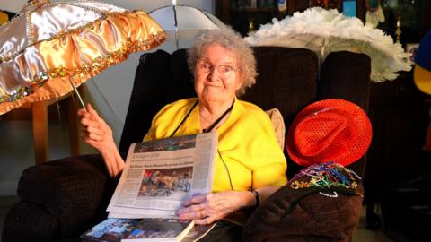 A woman smiles at the camera. She is sat on a brown armchair and hold a newspaper up to the camera. She is also holding a gold umbrella.