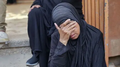 A woman sits on a stairway with her hand on her face, appearing distressed