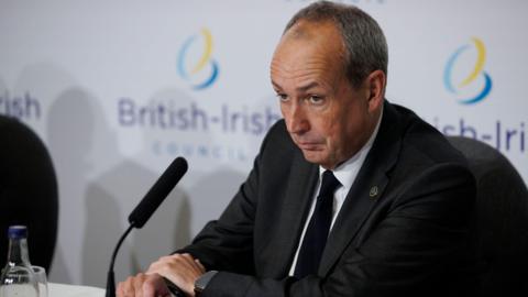 Jersey Chief Minister Lyndon Farnham sat wearing a black suit and tie with a white shirt sat in front of a microphone at a long table. The British-Irish Council logo can be seen on the screen behind him.