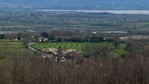 A photo showing Tait's Hill beauty spot near Stinchcombe. The photo is taken from a hill looking down on a circular area of green land surrounded by homes. In the background is the River Severn.