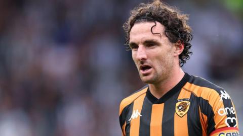 Lewie Coyle playing football for Hull City. He is wearing a traditional black and amber striped football shirt featuring the club's Tiger crest. He has long curly brown hair and is also wearing a rainbow-striped captain's armband.